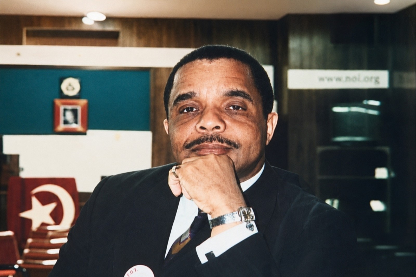 Brother Munir Muhammad in the C.R.O.E. studio, seated with chin resting on his hand, the Nation of Islam flag visible behind him. A photograph from the era of the recording.